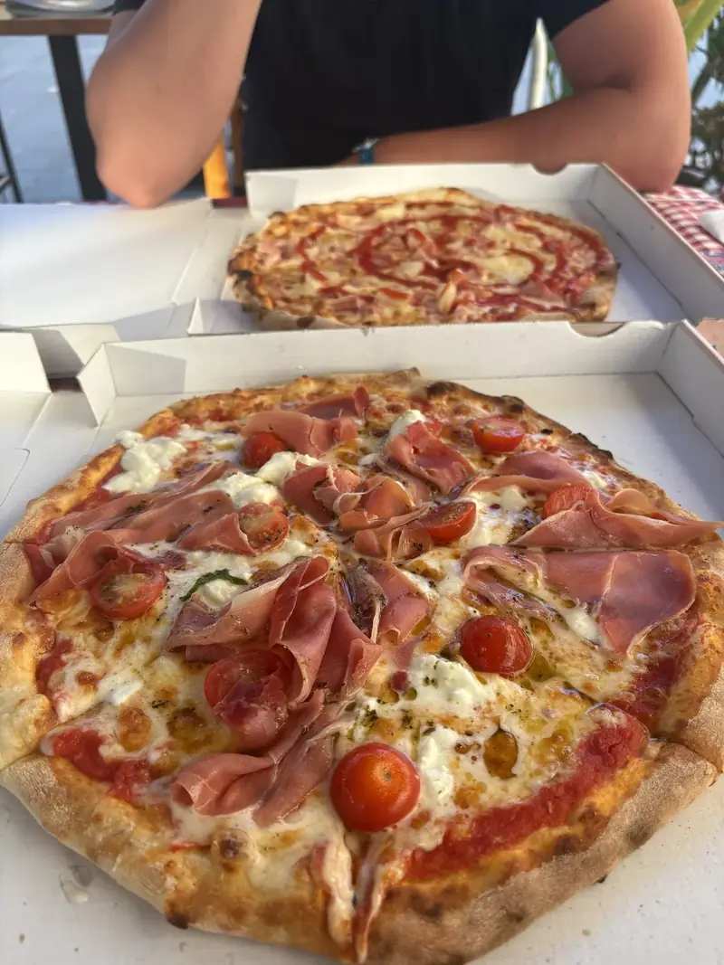 Dos pizzas en cajas de cartón abiertas sobre la mesa de la terraza, la de delante de prosciutto crudo con mozzarella y tomate cherry