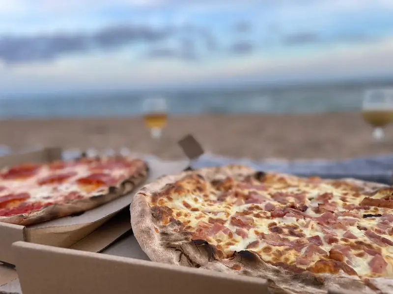 Dos pizzas en cajas de cartón abiertas sobre la arena de la playa de Pineda de Mar, con el mar al fondo y dos cañas de cerveza